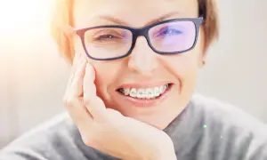 Uśmiechnięta kobieta w okularach, z aparatem na zębach, delikatnie dotyka policzka. Może odczuwać lekki ból zębów po założeniu aparatu.