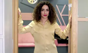 Helya Riazat, med lockigt mörkt hår och rött läppstift, står i en dörröppning i en guldig blus.