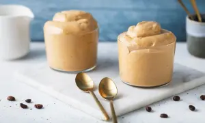 Due bicchieri di cremosa schiuma di caffè, perfetta per la tua ricetta crema di caffè senza panna. Due cucchiaini dorati e chicchi di caffè completano la scena.