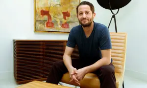 Ian Kirkpatrick, a man with a beard and a blue t-shirt, sits in a tan leather chair in a modern room with abstract art.