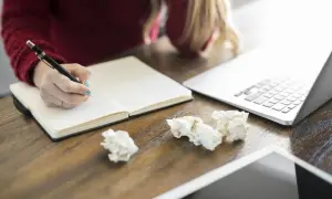 Dłoń pisząca w notatniku obok laptopa i zmiętych kartek.