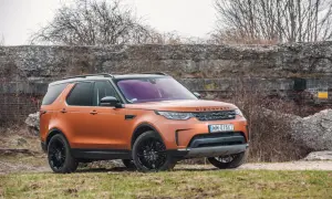Pomarańczowy Land Rover Discovery 5 z czarnymi felgami stoi na trawiastym podłożu, na tle kamiennego muru i suchych krzewów.