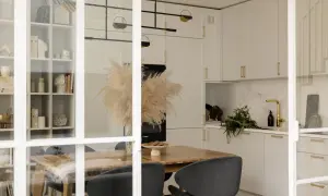 Elegancka kuchnia z białymi frontami i uchwytami w kolorze mosiądz. W centralnym punkcie stół z suszonymi trawami i czarnymi krzesłami.