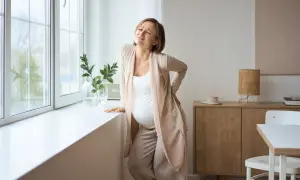 Gravid kvinna med ont i svanskotan lutar sig mot ett fönster. Hon håller handen på ryggen och grimaserar av smärta.