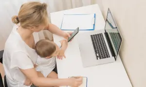 Kobieta z dzieckiem na kolanach sprawdza telefon, obok laptop i notatnik. Planuje wnioski o zasiłek macierzyński.