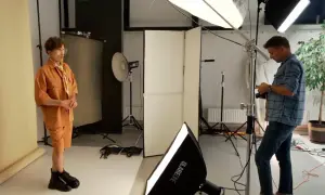 Model w pomarańczowym stroju pozuje w studiu, gdzie fotograf ustawia sprzęt, tworząc idealne naturalne źródła światła.