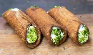 Trzy chrupiące włoskie słodycze, cannoli, wypełnione kremem i posypane pistacjami, leżą na drewnianej desce.