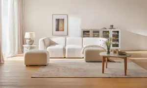 Przytulny salon z kanapą i pufą w odcieniach beżu farby. Jasne zasłony, drewniana podłoga i subtelne dekoracje tworzą spokojną atmosferę.