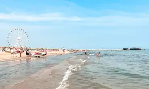 Słoneczny dzień na plaży w Rimini. Ludzie spacerują, bawią się w wodzie, a w oddali widać diabelski młyn. Idealne miejsce na wakacje!