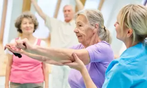 Seniorzy wykonują ćwiczenia na alzheimera z fizjoterapeutką. W tle inni uczestnicy ćwiczą z hantlami.