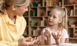 Kobieta i dziewczynka rozmawiają, gestykulując. W tle regały z książkami.