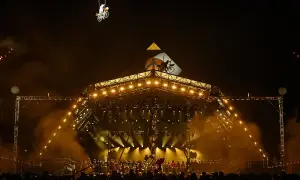 A performer on a bicycle is suspended in the air above a brightly lit stage at Glastonbury. The crowd below anticipates the next act, checking their Glastonbury set times.