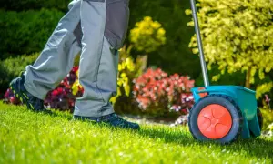 Osoba w szarych spodniach i czarnych butach z zielonymi akcentami wykonuje dosiewanie trawy za pomocą zielonego rozsiewacza.