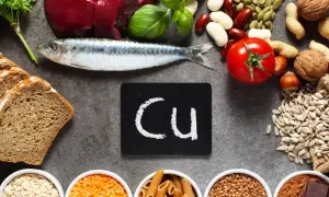 Na stole leżą ryba, chleb, pomidor, orzechy, nasiona i inne produkty bogate w miedź. Tabliczka z napisem "Cu" symbolizuje ten pierwiastek.