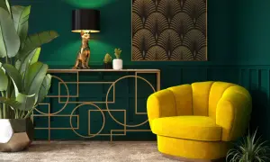 Elegancka tkanina art deco zdobi ścianę nad złotą konsolą z lampą w kształcie kota. Obok żółty fotel i bujna roślina.