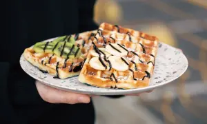 Gofry z bananem i kiwi, polane czekoladą, posypane cukrem perlistym. Pyszny deser na talerzu.