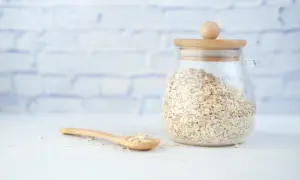 Szklany słoik pełen płatków owsianych ig, obok drewniana łyżka z kilkoma płatkami.