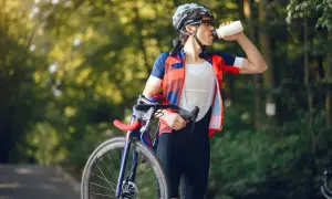 Kolarz w stroju sportowym pije wodę z bidonu, dbając o odpowiednią dietę kolarza. W tle zielony las.