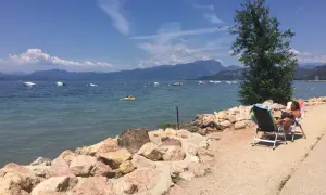 Una donna legge su una sedia a sdraio sulla riva rocciosa di Cisano, Gardasee. Barche galleggiano sull'acqua calma con montagne sullo sfondo.