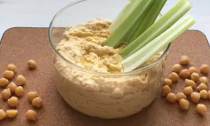 Pyszna pasta z ciecierzycy w szklanej misce, podana z chrupiącymi łodygami selera i ziarnami ciecierzycy. Idealna przekąska!