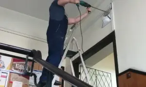 Mężczyzna w kombinezonie wierci w ścianie, stojąc na drabinie bhp.