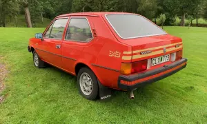 En röd Volvo 343 står på en grön gräsmatta med skog i bakgrunden. Bilen har en gul rand längs sidan och texten "343" i guld.