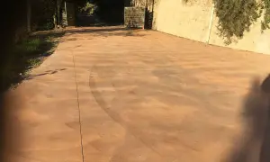 Un camino de hormigón de colores, con marcas de neumáticos, se extiende hacia una entrada flanqueada por vegetación y un muro de piedra.