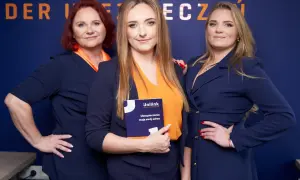 Trzy kobiety w granatowych marynarkach i pomarańczowych bluzkach pozują na tle niebieskiej ściany z logo.