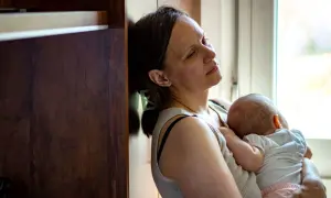 Zmęczona matka tuli śpiące dziecko, opierając się o mebel. Widać jej wyraz twarzy, który sugeruje wypalenie rodzicielskie.