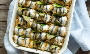 Sarde a beccafico pronte per essere infornate, disposte in una teglia con foglie di alloro e panatura dorata.