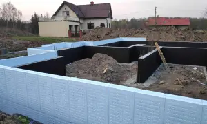 Budowa domu w toku. Widoczna izolacja fundamentów z użyciem płyt XPS. W tle dom jednorodzinny.