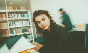Młoda kobieta w okularach siedzi przy biurku w bibliotece, otoczona książkami.