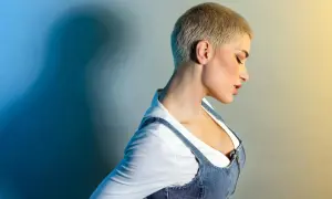 Kobieta z **buzz cut blond** w dżinsowej sukience na szelkach i białej bluzce. Ciemny cień na niebieskim tle.