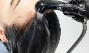 Kobieta z ciemnymi włosami poddaje się zabiegowi **reverse washing**. Woda spływa po jej głowie, a dłoń w czarnej rękawiczce trzyma słuchawkę prysznicową.