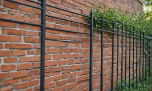 Czerwone, ceglane ogrodzenie z czarnymi, metalowymi prętami i zielonymi pnączami.