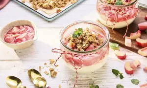Zwei Gläser mit Rhabarber Crumble, garniert mit Minze. Frischer Rhabarber liegt daneben.