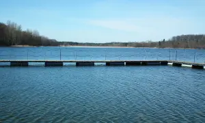 Drewniane molo na spokojnym jeziorze Pobiedziska, otoczonym lasem. Woda lekko faluje, niebo błękitne.