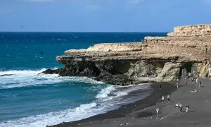 Fuerteventura Urlaub Tipps: Schwarzer Strand mit Felsen und türkisblauem Meer. Menschen spazieren und entspannen.