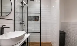 Moderno murete baño con ducha de cristal, lavabo blanco y grifería negra. Azulejos blancos y suelo de madera.