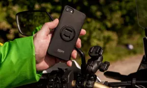 Dłoń trzyma czarne etui na telefon z mocowaniem SP Connect, obok uchwytu na kierownicy motocykla.
