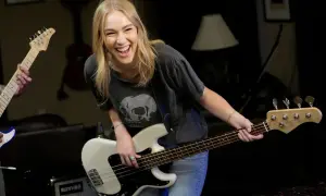 Uśmiechnięta blondynka gra na białym bas instrumencie, w tle inny gitarzysta.