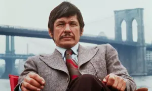 Charles Bronson w garniturze, z wąsami, siedzi przed mostem Brooklińskim. Ikona kina akcji, znany z wielu filmów.