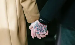 Manos entrelazadas, una con un tatuaje de rosa en fase lechosa, la otra envuelta en un abrigo camel.