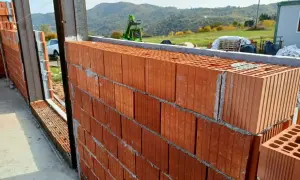 Muro in costruzione con blocchi di laterizio, un materiale che resiste al fuoco, con vista su colline verdi e un escavatore.