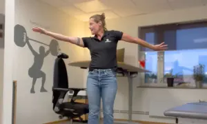 Kvinna utför en rörelse för att stretcha axel. Hon står framför en väggmålning av en tyngdlyftare.
