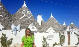 Una donna sorride davanti ai trulli di Alberobello, patrimonio UNESCO, con un grande fiocco azzurro appeso a un cono.