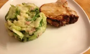 Ein gebratenes Schweinekotelett liegt neben einem Berg aus Kartoffelpüree mit Speck und **endivien untereinander**.