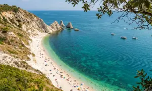 Spiaggia affollata ai piedi del Monte Conero, con acque cristalline e barche all'orizzonte.