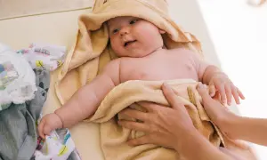 Söt bebis i handduk, kanske med tyst reflux bebis symtom. Bredvid ligger kläder och blöjor.