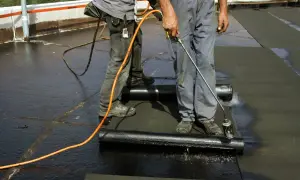 Zwei Dachdecker arbeiten auf einem Flachdach. Einer hält einen Brenner, um die Dachpappe zu schweißen.